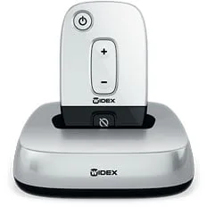 A Widex TV-DEX sits in its charging dock—a white, rectangular device with a power button, volume control, and the Widex logo—arranged on a sleek silver base.